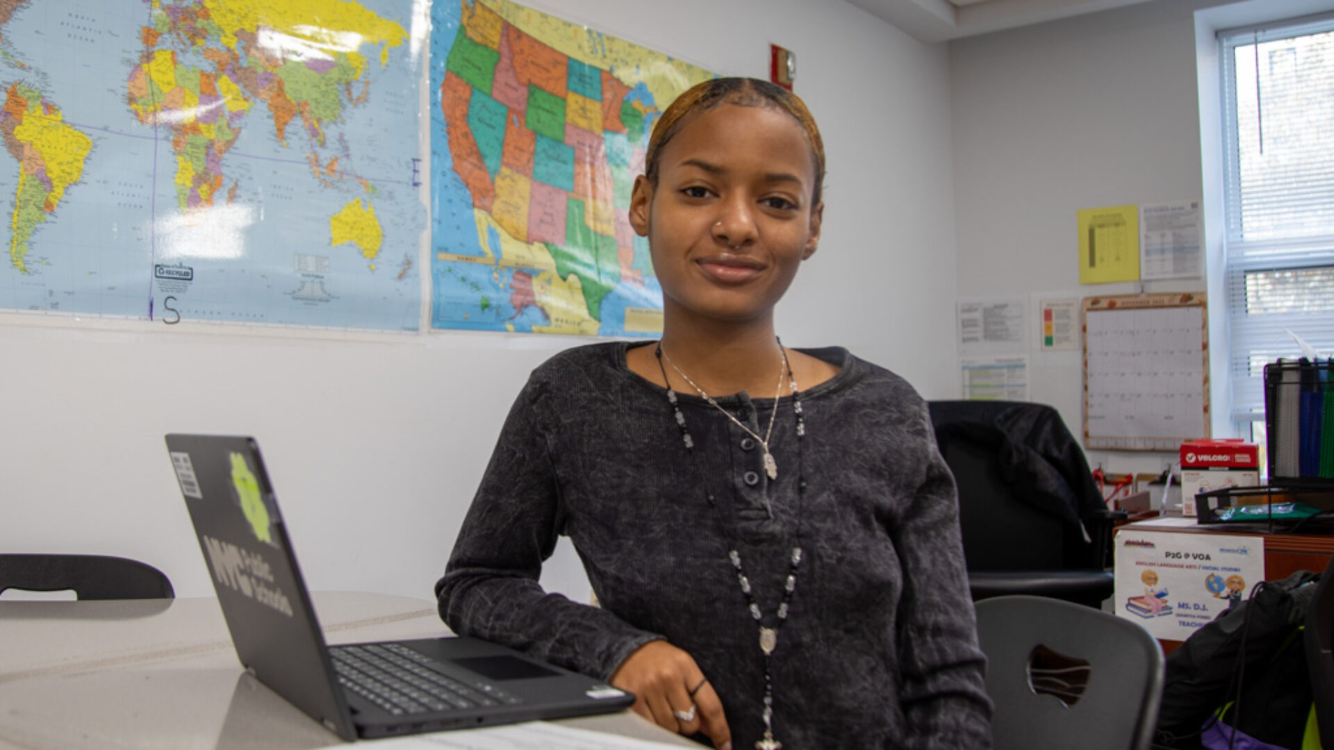 A young woman smiles at the camera while seated at a table with an open laptop.