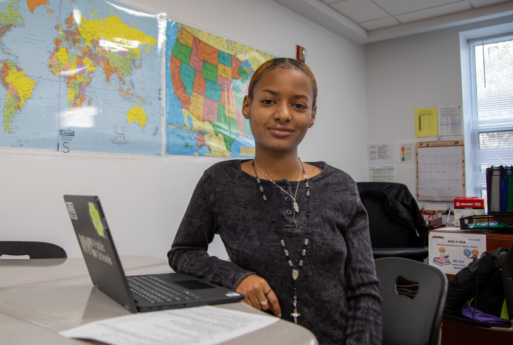 A young woman smiles at the camera while seated at a table with an open laptop.