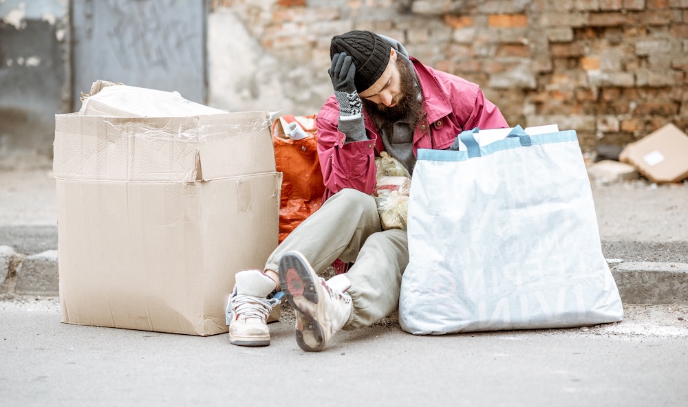 Man with winter gloves and hat sitting on ground with a box and bags beside him.