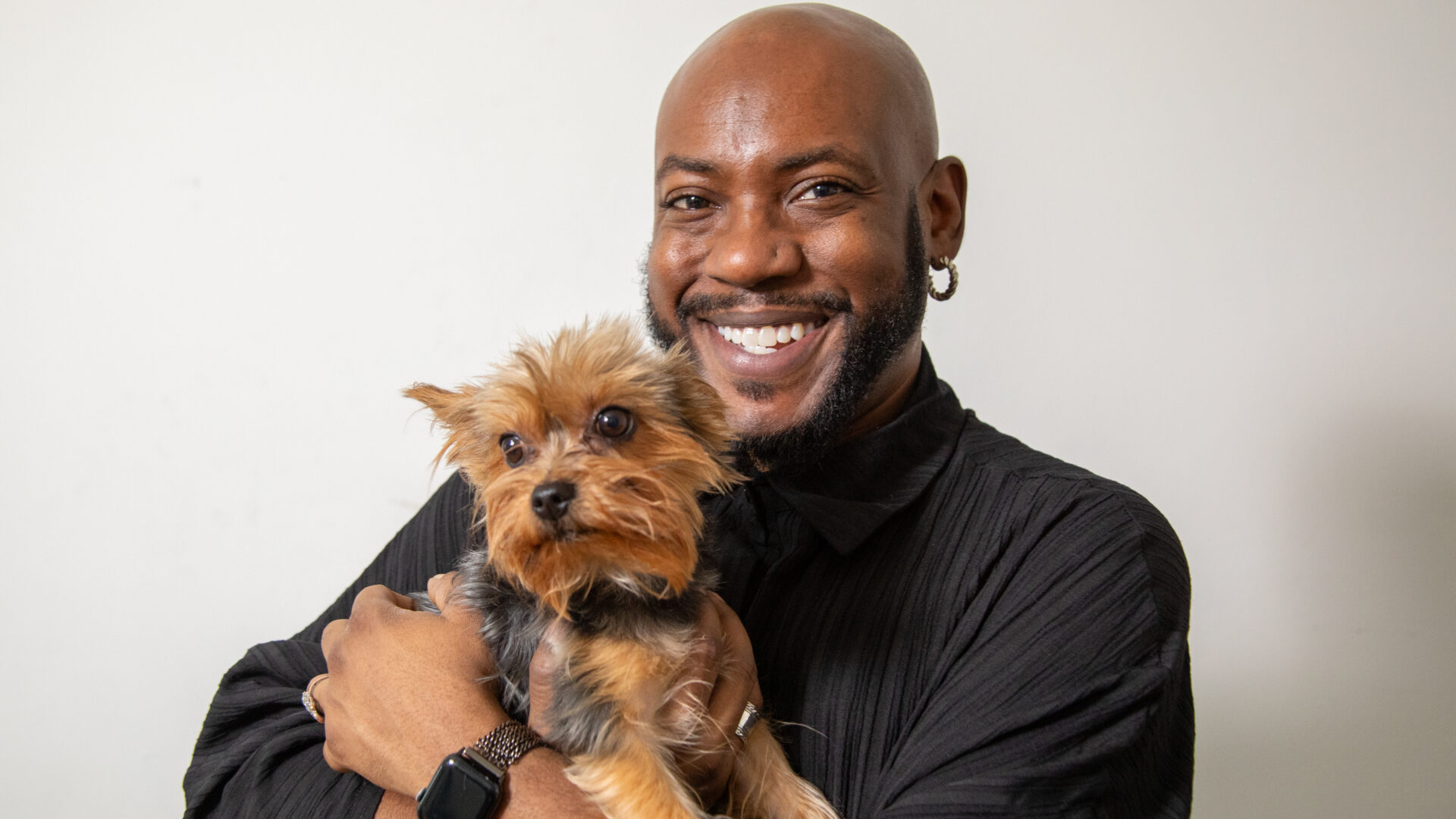 A smiling, Black man carrying a small dog against a white background.
