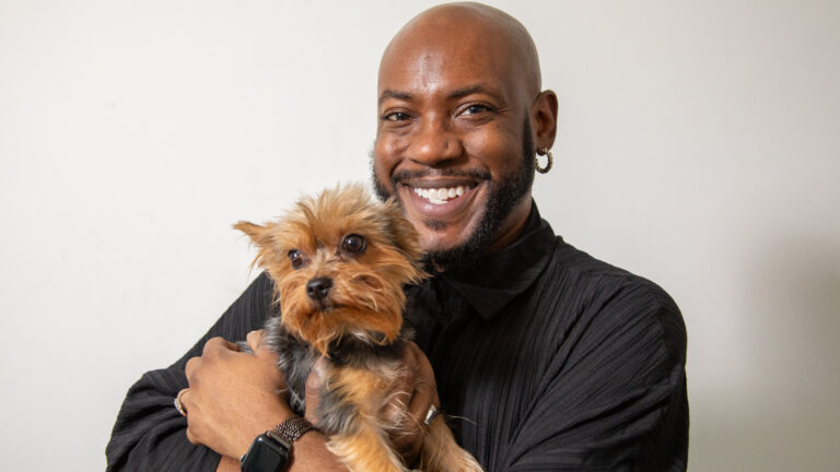 A smiling, Black man carrying a small dog against a white background.