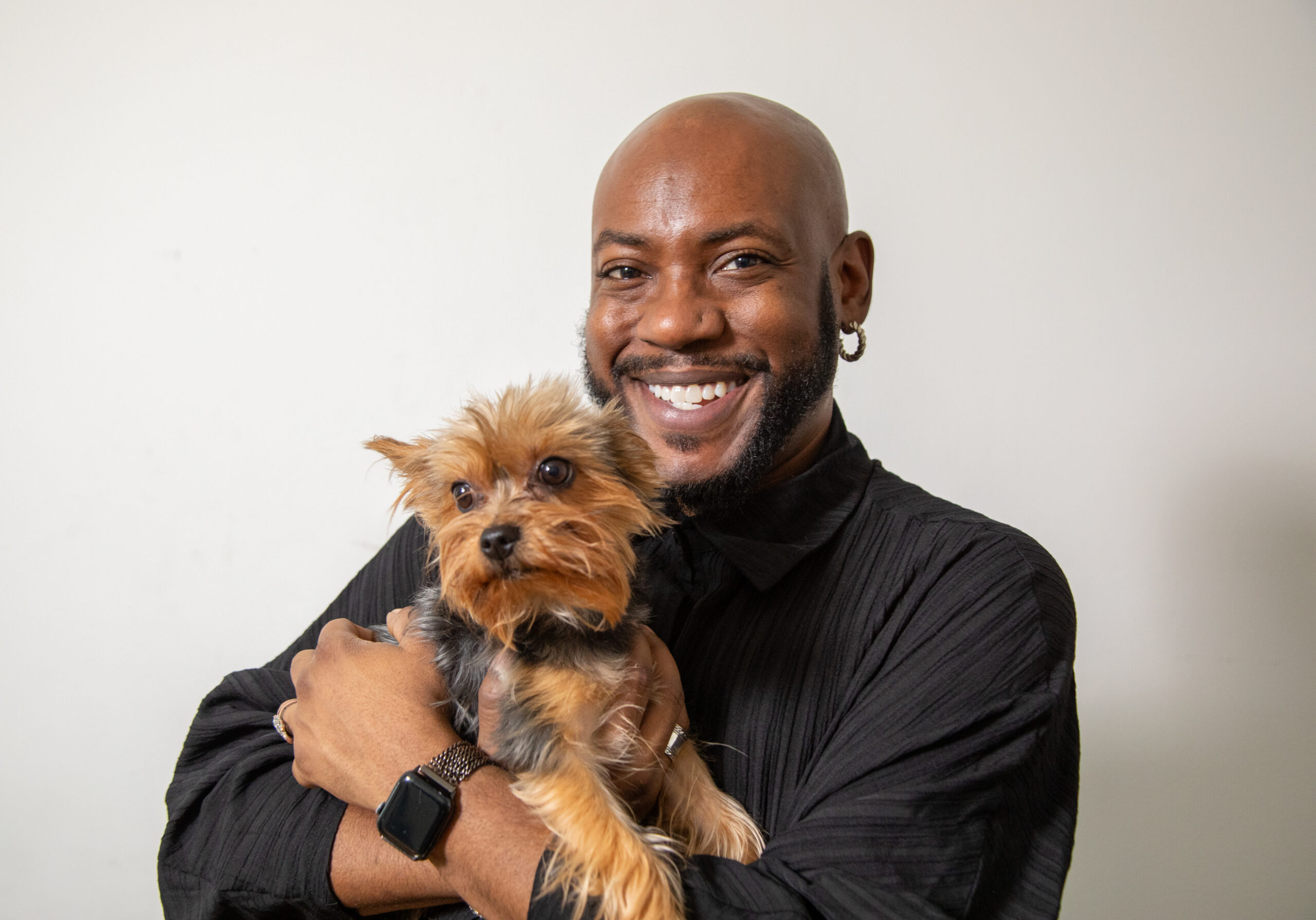 A smiling, Black man carrying a small dog against a white background.