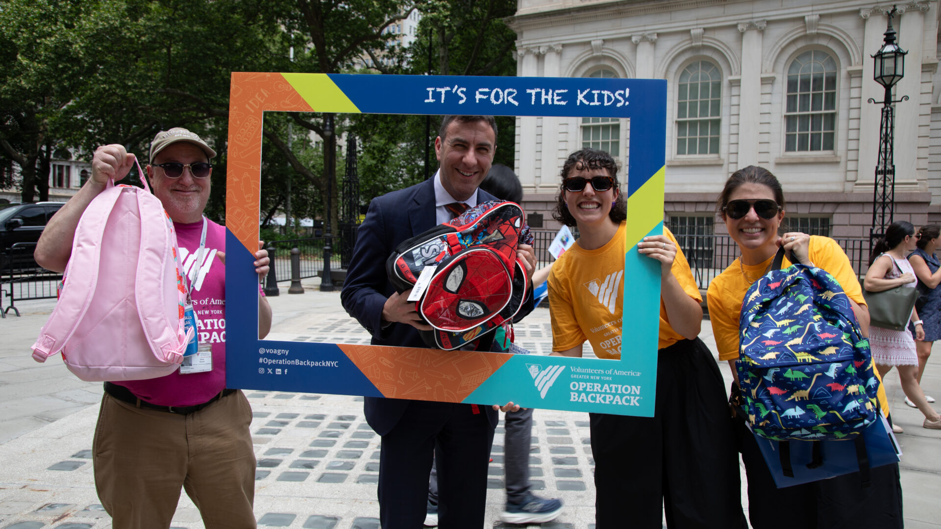 VOA-GNY staff stand with Councilmember Eric Dinowitz holding an Operation Backpack frame and children's backpacks outside City Hall