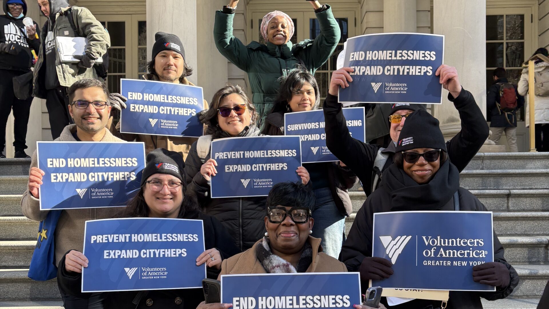 Group of eight staff members hold signs reading "End homelessness, expand CityFHEPS" outside City Hall.