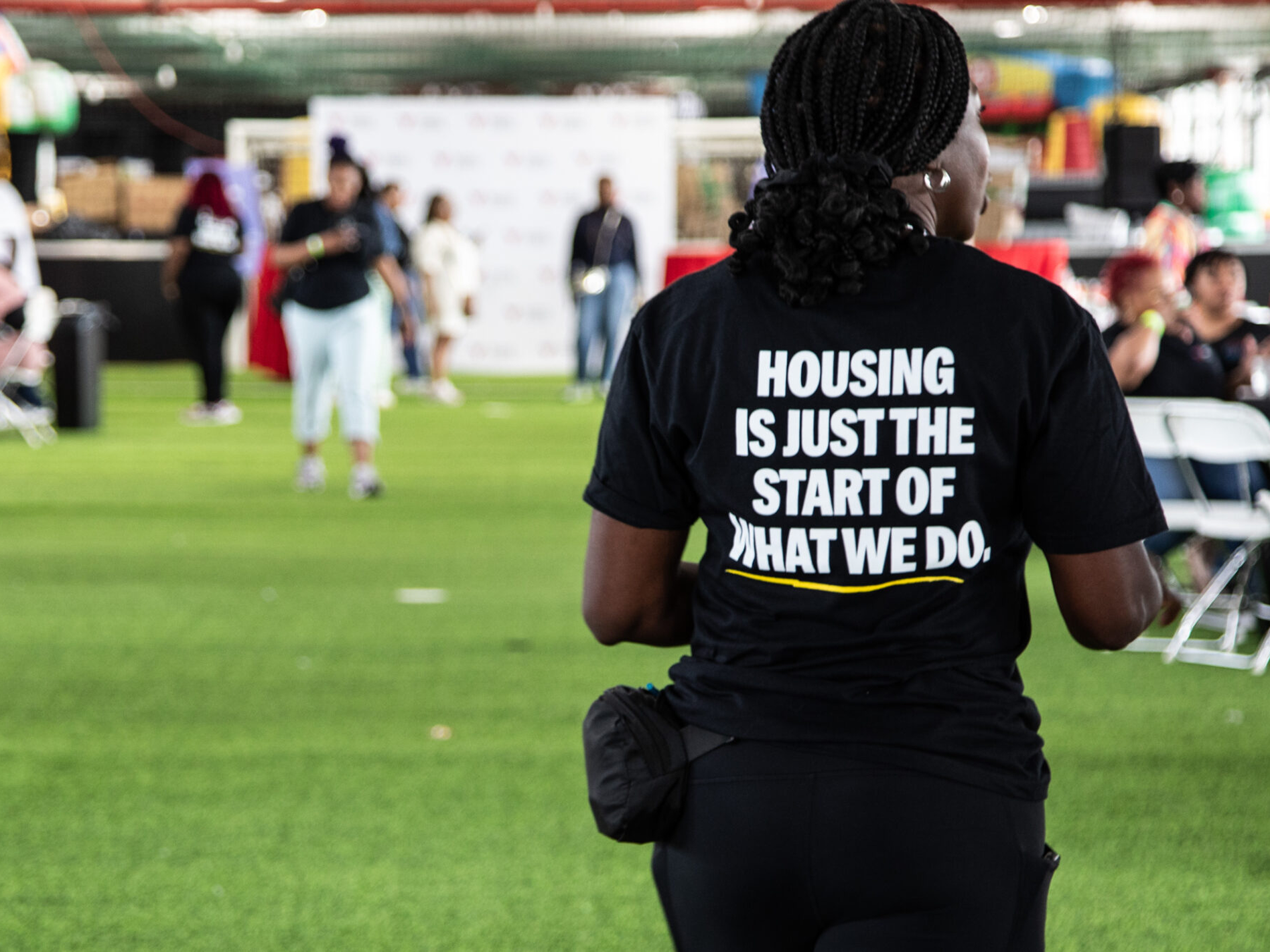 Person walking at an outdoor event while wearing a shirt featuring the text "Housing is just the start of what we do"