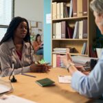 Young adult Black woman sitting across from middle aged Caucasian female social worker in office discussing social services assistance, paperwork and identification documents visible on desk