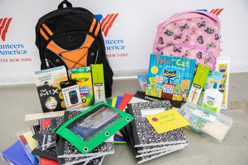 School supplies piled in front of two backpacks against a background with a logo for Volunteers of America-Greater New York.