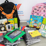 School supplies piled in front of two backpacks against a background with a logo for Volunteers of America-Greater New York.